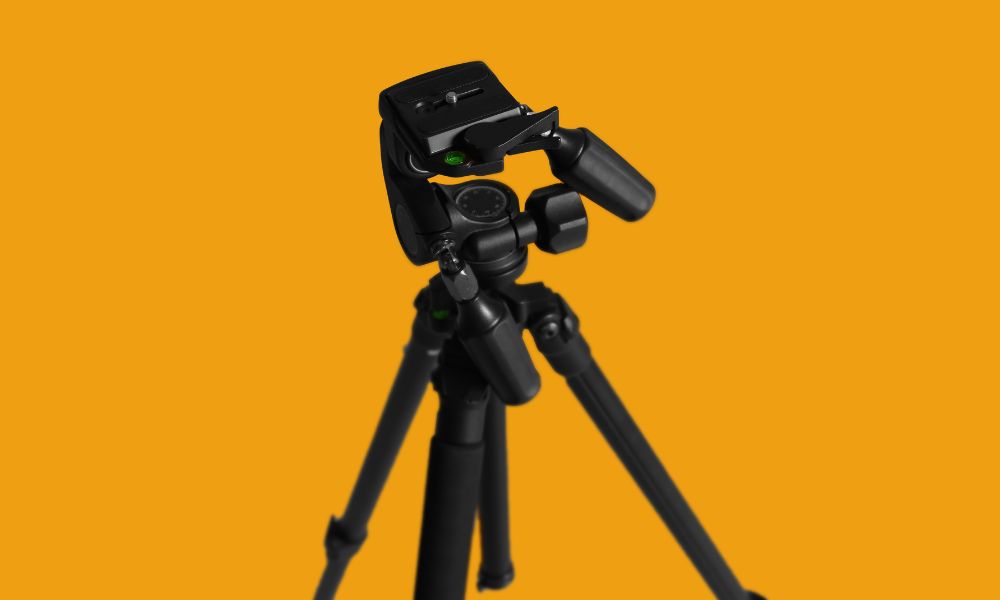 A black camera tripod with a pan-and-tilt head is positioned against a solid orange background, with its legs extended