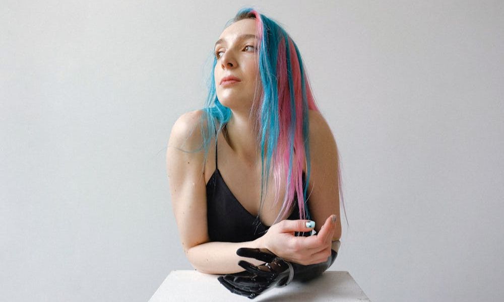 A woman with long, vibrant pink and blue hair, wearing a black spaghetti strap top, leans forward with one arm on a white surface and a prosthetic hand visible. She is looking thoughtfully to her left against a plain white background