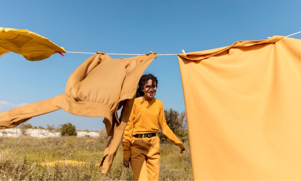 A young woman with dark, curly hair and light skin, wearing a mustard yellow long-sleeved shirt and pants, stands smiling in a grassy field with yellow garments hanging on a clothesline, under a clear blue sky