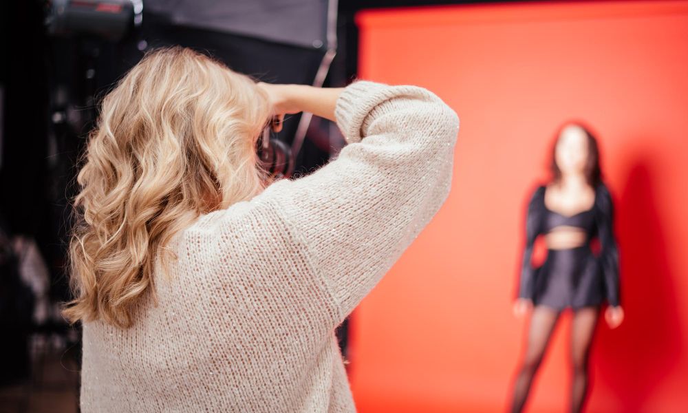 A blonde female photographer, seen from behind, is holding a camera up to her eye, photographing a blurred model in a black outfit against a vibrant red backdrop in a studio setting