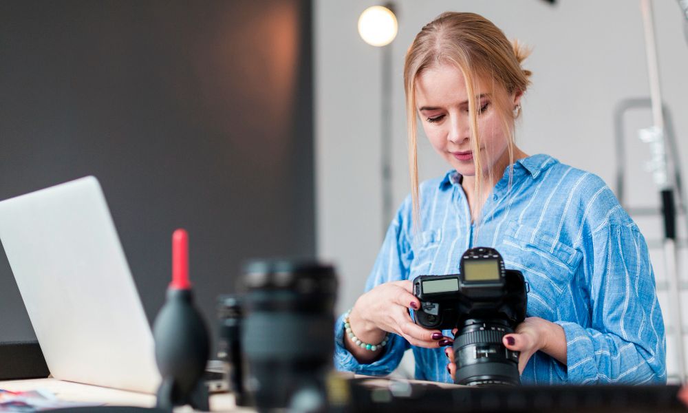 A woman with light brown hair, wearing a blue striped shirt, is looking down and adjusting the settings on a black DSLR camera