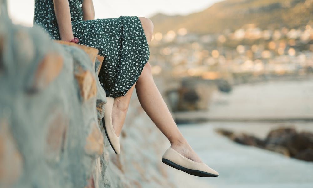 A female in a dark green floral dress with bare legs sits on a stone wall, wearing light-colored flat shoes, with a blurred coastal landscape in the background