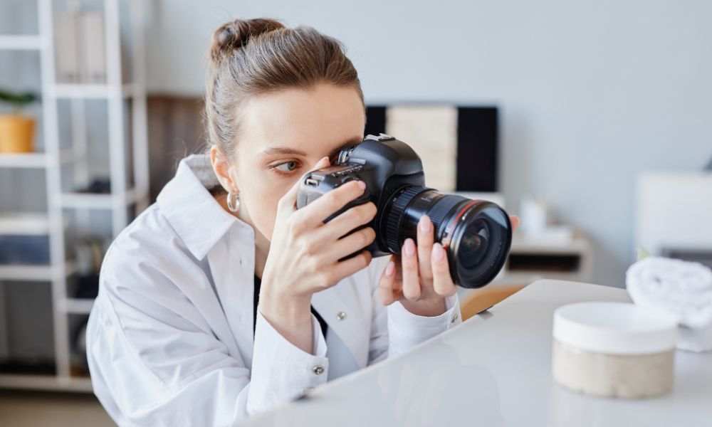 A woman with her hair in a bun and wearing a white collared shirt leans over a light-colored surface, looking through the viewfinder of a black DSLR camera, with a small white jar of product in the foreground