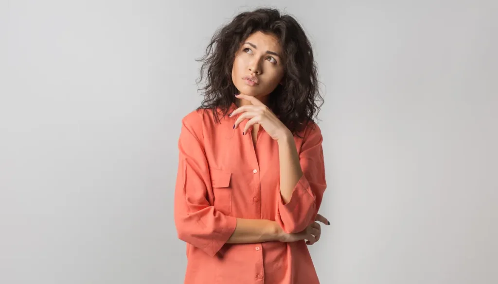 A young woman with curly brown hair and an orange shirt looks upwards and to the right with a thoughtful expression, her hand touching her chin, against a plain light gray background