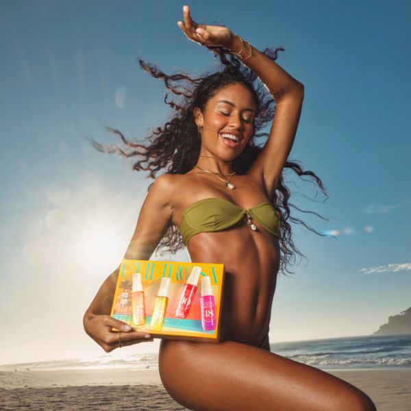 Joyful woman with curly hair in a green bikini, holding an orange box of colorful spray bottles on a sunny beach with ocean and sky in the background