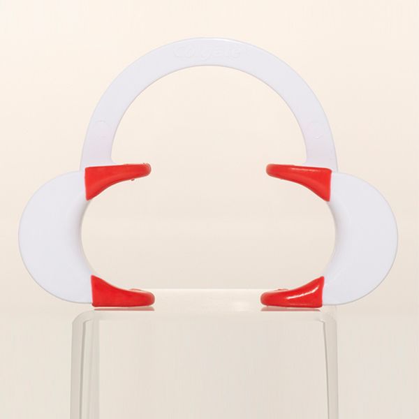 A white dental retractor, with red grips on the lower crescent-shaped parts, is positioned on a clear, reflective surface. The background is a plain, light beige color.