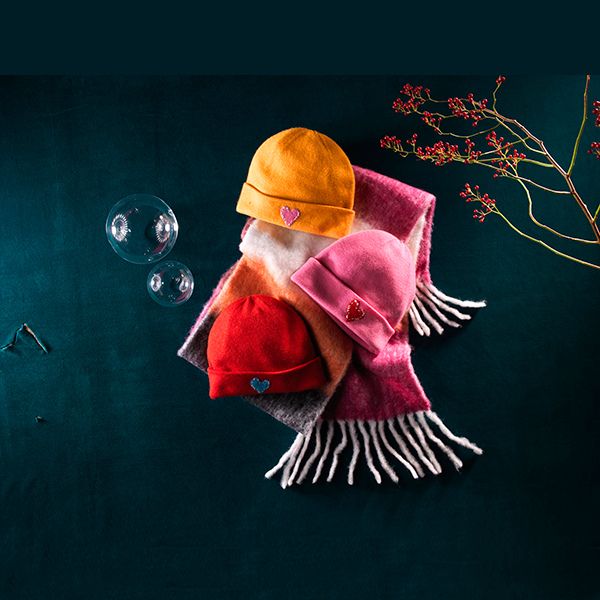 Three heart-patched beanies (orange, pink, red) on a striped scarf with bubbles and red berries on dark teal.