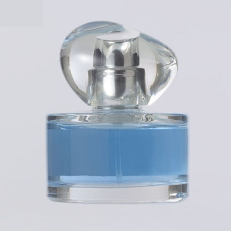 A round glass perfume bottle with a clear, sculpted cap, filled with light blue liquid, against a plain white background