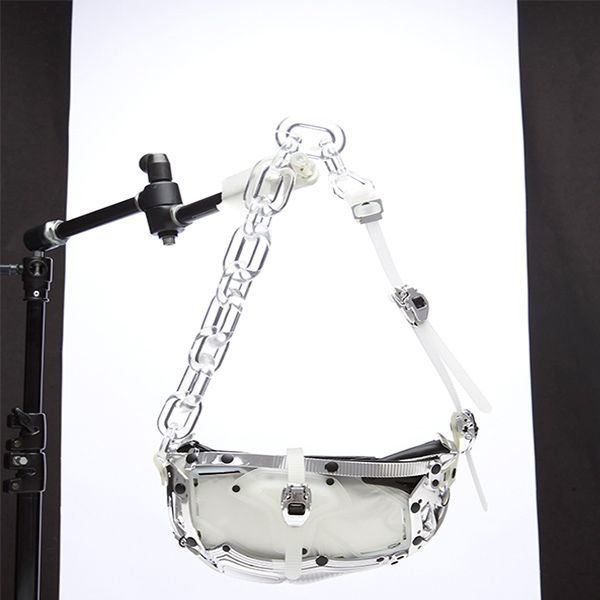 A transparent and white handbag with a chunky clear chain strap is suspended by visible white straps and a black support arm with clamps, against a pure white backdrop.