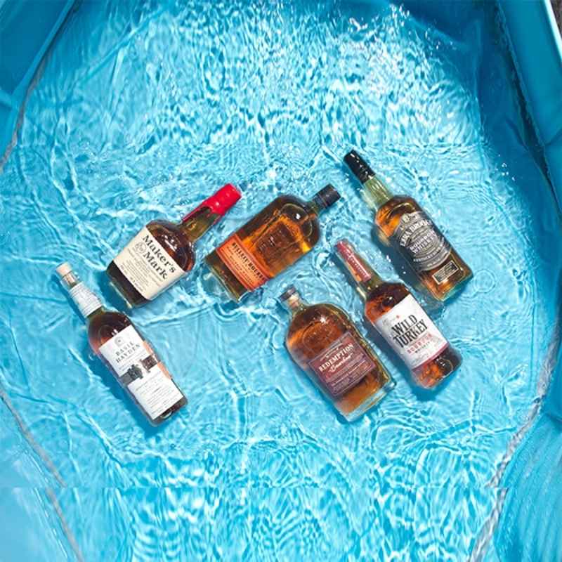 Six bottles of spirits, including Maker's Mark, Wild Turkey, and Redemption, float in clear blue water in a pool