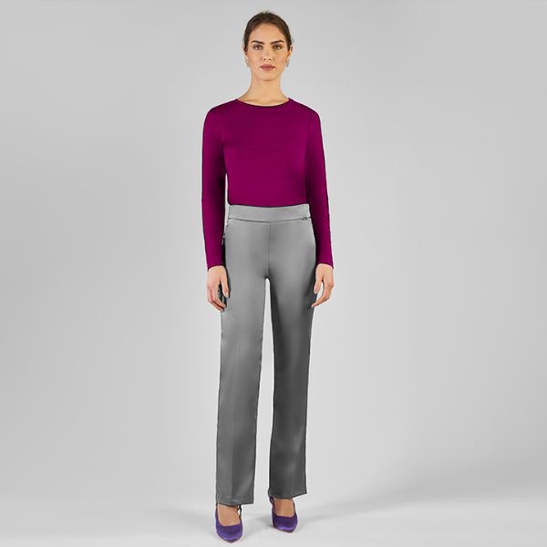 A full-length shot of a woman with dark hair pulled back, wearing a magenta long-sleeved top, shiny grey trousers, and purple high-heeled shoes, standing against a plain light grey background