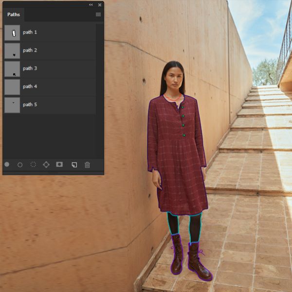 A woman in a long-sleeved maroon plaid dress, dark tights, and brown boots stands against a textured light brown wall and stairs. Overlaid on the image is a "Paths" panel from an editing software, showing outlines around parts of her clothing and shoes, indicating multiple clipping paths