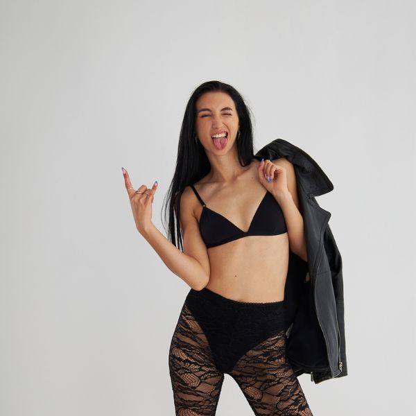 A woman in a black bralette and lace leggings playfully poses with her tongue out and one hand forming a rock gesture, holding a black leather jacket over her shoulder.