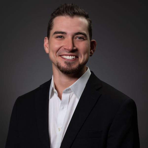 A smiling man in a black suit jacket and white shirt poses against a dark gray background in a professional headshot