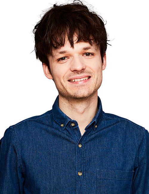 A smiling person with dark, slightly disheveled hair and a light beard wears a denim shirt, against a pure white background