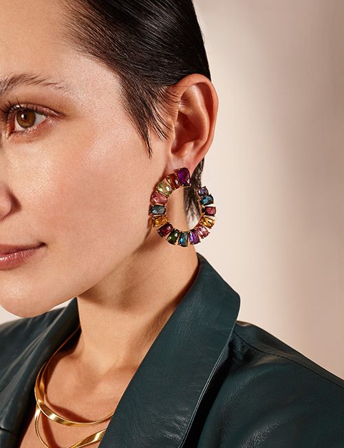 Retouched image of a close-up of a woman's face from the nose up, focusing on a large, gold hoop earring adorned with multi-colored gemstones