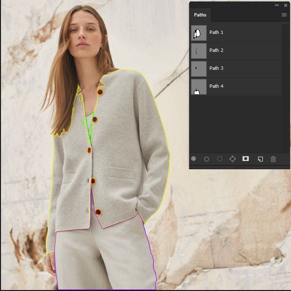 A young woman wearing a light grey button-up cardigan over a white top and light-colored trousers stands against a textured light-colored wall. Overlaid on the image is a "Paths" panel from an editing software, with colored outlines around her clothing and body, indicating multiple clipping paths