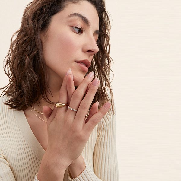 A woman with wavy brown hair poses with her hands gently clasped near her face, wearing a cream ribbed top and accessorized with gold and diamond rings and layered necklaces