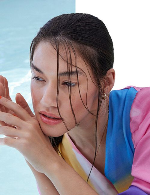 A woman with dark, wet hair, possibly just out of water, looks to the side with a pensive expression. She appears to be wearing a colorful, tie-dye style top. The background is split, showing a pale blue, possibly water, on the left and white on the right.
