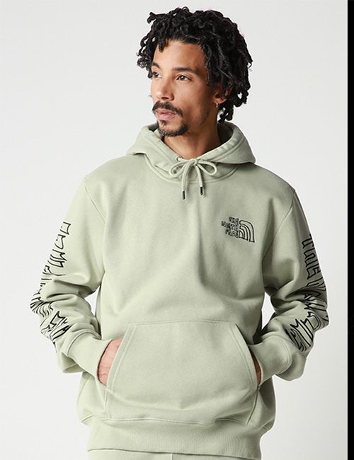 A person with dark, textured hair and a beard wears a light green "The North Face" hoodie with graphic designs on the sleeves, standing with hands in the front pocket against a white background.