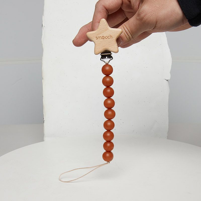 A hand holds up a baby pacifier clip, featuring a wooden star-shaped clasp engraved with "smooch" and a chain of nine reddish-brown silicone beads.