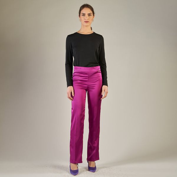 A full-length shot of a woman with dark hair pulled back, wearing a black long-sleeved top, shiny fuchsia pink trousers, and purple high-heeled shoes, standing against a plain light brown or grey background