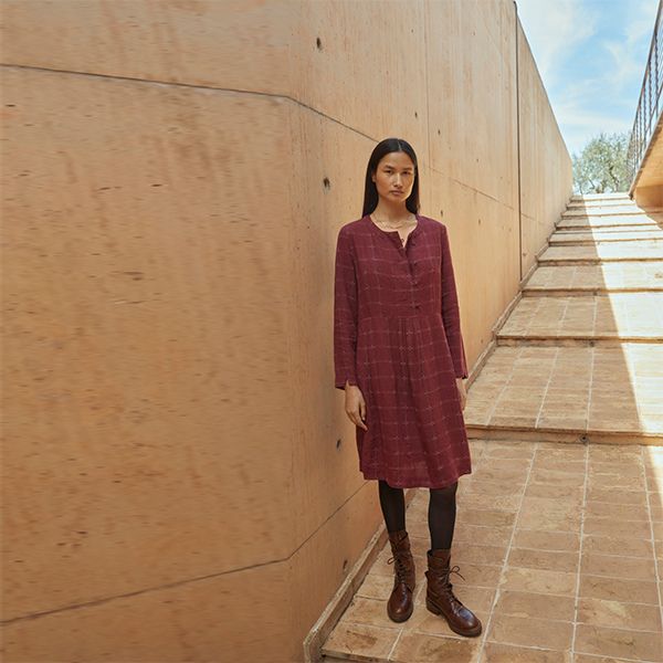 A woman with long dark hair, wearing a long-sleeved maroon plaid dress, dark tights, and brown boots, stands against a textured light brown wall next to a set of stairs