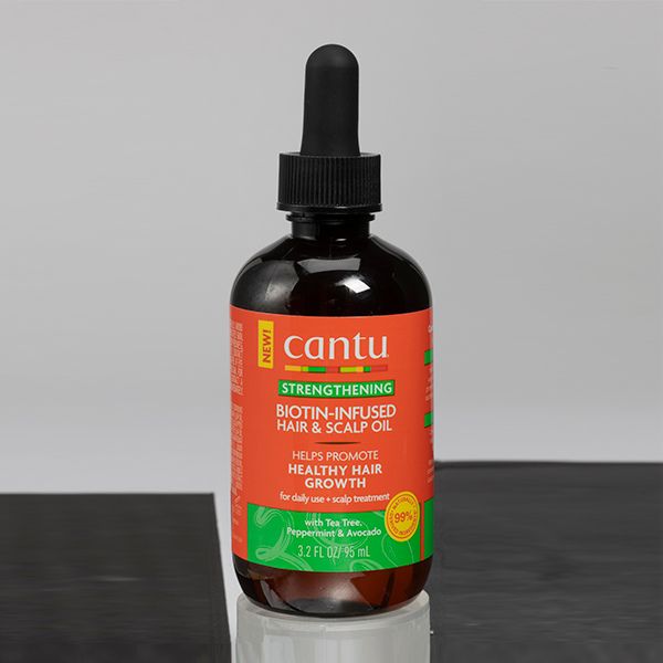 A bottle of Cantu Strengthening Biotin-Infused Hair & Scalp Oil, 3.2 fl oz / 95 ml, with a black dropper top and an orange and green label, sitting on a reflective black and white surface