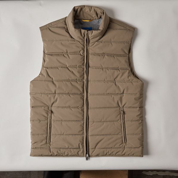 A khaki-colored puffer vest with a high collar and a front zipper, laid flat on a light-colored surface with some wrinkles and shadows.