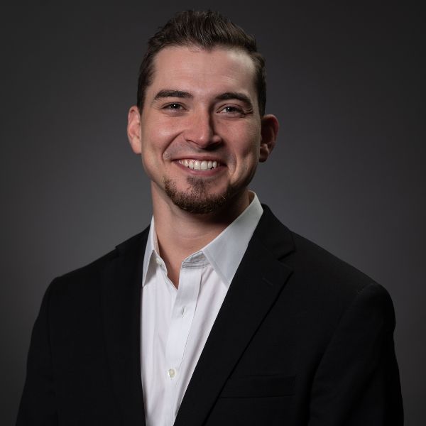 A smiling man with a beard and slicked-back dark hair, wearing a black suit jacket over a white collared shirt, against a dark grey background