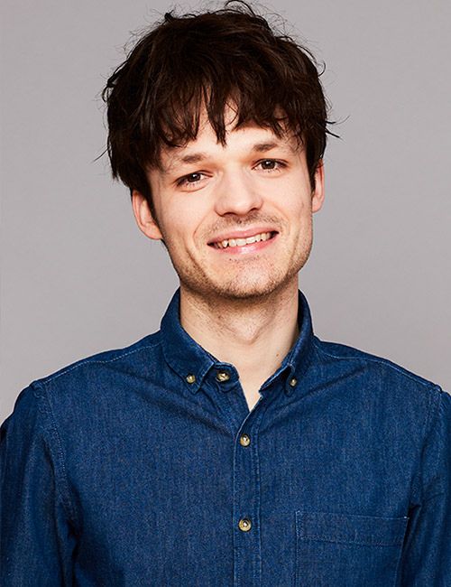 A smiling person with dark, slightly disheveled hair and a light beard wears a denim shirt, against a plain grey background