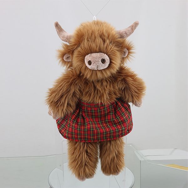 A fluffy brown Highland cow plush toy wearing a red plaid kilt is suspended, likely by a string, against a light grey background with subtle reflections on a clear surface below