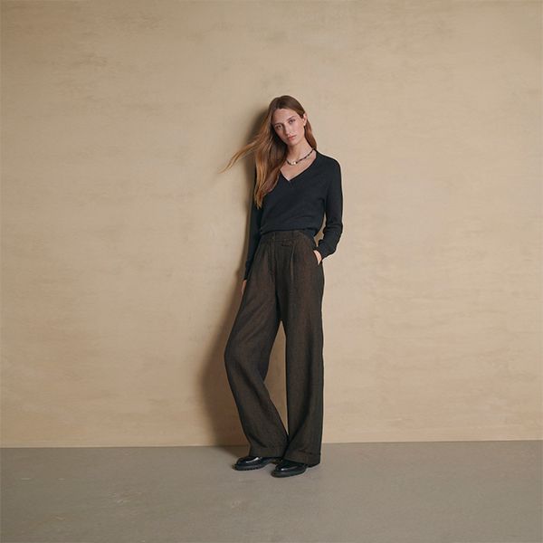 An attractive woman with long, light brown hair, wearing a dark V-neck long-sleeve top, dark brown wide-leg trousers, and black shoes, stands casually with one hand in her pocket against a beige wall and grey floor