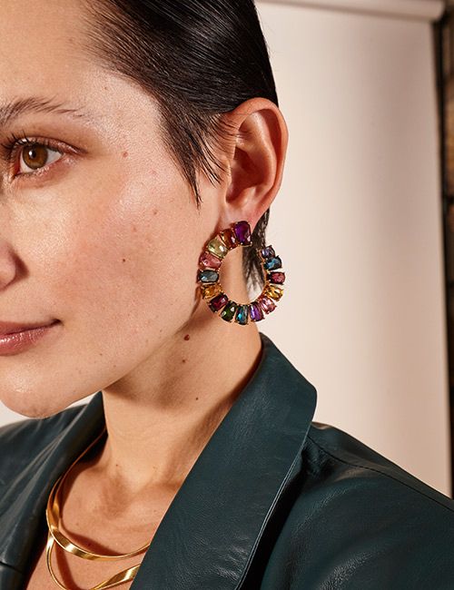 A close-up of a woman's face from the nose up, focusing on a large, gold hoop earring adorned with multi-colored gemstones