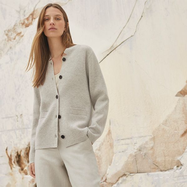 A young woman with long, light brown hair, wearing a light grey button-up cardigan over a white top and light-colored trousers, stands against a textured light-colored wall