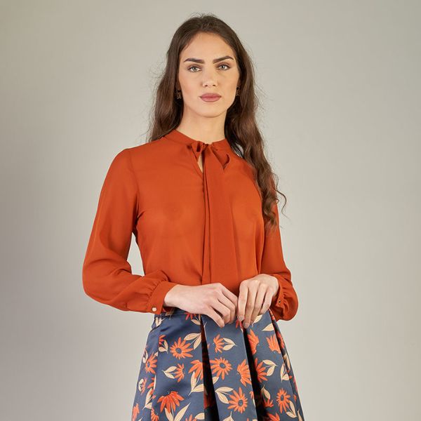 A woman with long, wavy brown hair wears an orange blouse with a tie-neck detail and a dark blue skirt with an orange floral pattern