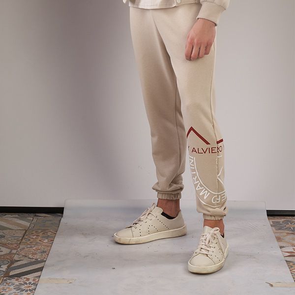 person wearing beige sweatpants with red/white graphics and light sneakers stand on a rolled grey paper backdrop with patterned tiles visible