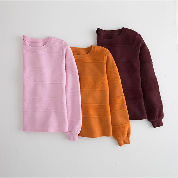 Three textured long-sleeved shirts, in shades of pink, orange, and maroon, are neatly laid out, slightly overlapping each other on a white surface