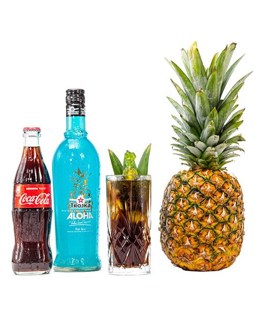 A product shot featuring a Coca-Cola bottle, a blue bottle of Trojka Aloha liqueur, a tall glass cocktail garnished with leaves, and a whole pineapple, all arranged on a white background.
