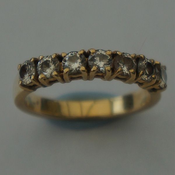 A slightly blurry close-up of a gold ring featuring seven round, clear gemstones, on a muted blue-grey background