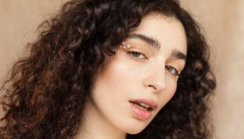 A close-up of a woman with dark, curly hair and light skin, looking slightly to her right. She has small pearls or beads near the outer corners of her eyes