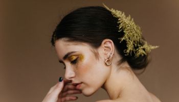 A side profile of a woman with dark hair styled in a bun, adorned with a gold leafy hairpiece. She has glittery gold eye makeup and a subtle blush, with a gold hoop earring visible