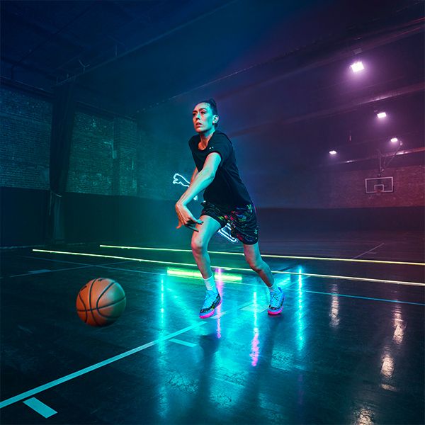 A female basketball player in a black t-shirt and colorful shorts is mid-dribble on a court lit by vibrant pink, blue, and green neon lights