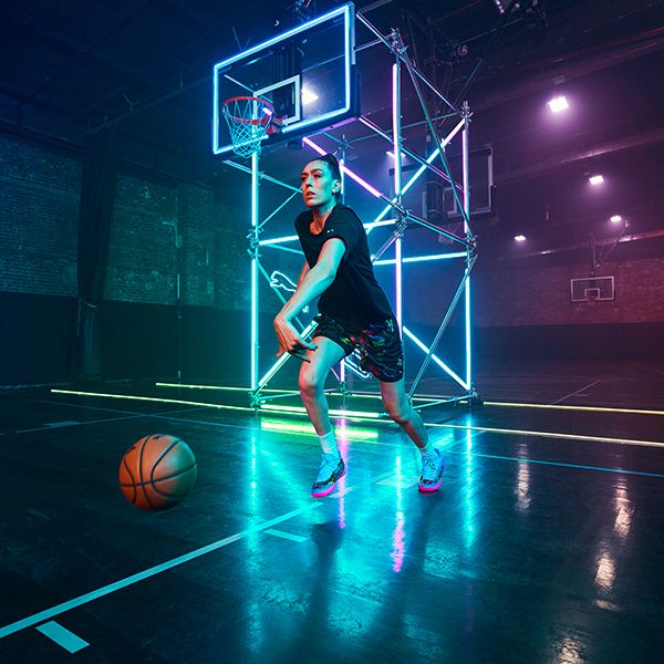 A female basketball player in a black t-shirt and colorful shorts is mid-dribble on a court lit by vibrant pink, blue, and green neon lights, with a basketball hoop in the background