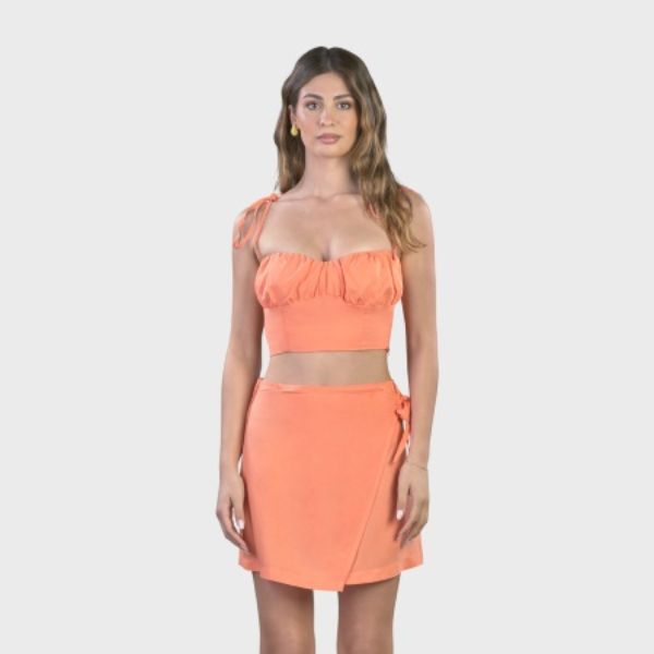 A woman with long brown hair, wearing an orange two-piece outfit consisting of a cropped camisole with shoulder ties and a wrap-style mini skirt, looking directly at the camera
